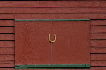A rusty horseshoe pointed upward on a red barn stable door with green trim. The lucky symbol is a...