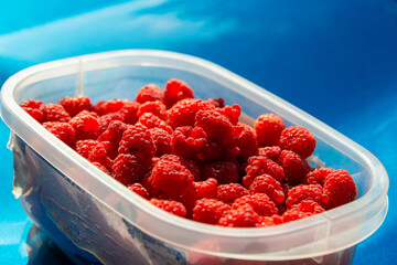 A plastic container of plump organic red wild raspberries. The berries are glossy with a sheen. The soft ripe clustered round sweet fruit are crimson red and textured. The basket is full.