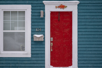 The exterior of a blue wooden cottage with a bright red solid metal door. There's a silver metal mailbox next to a closed glass window with a trim. A wooden fish hangs over the door.