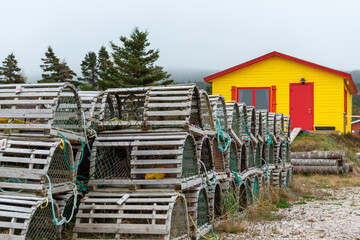 Rows of traditional wooden lobster pots and traps arestacked on a grassy meadow. The cages are made of small wood sticks, green nylon fishing rope. There's a yellow storage building with a red door.  