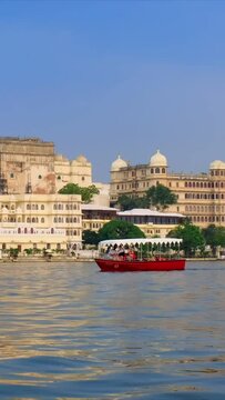 Udaipur City Palace on the bank of lake Pichola - Rajput architecture of Mewar dynasty rulers of Rajasthan. Tourist boat is heading to the side. Udaipur, India. Camera pan