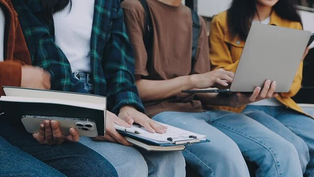 Group of happy young Asian college students sitting on a bench, looking at a laptop screen, discussing and brainstorming on their school project together.