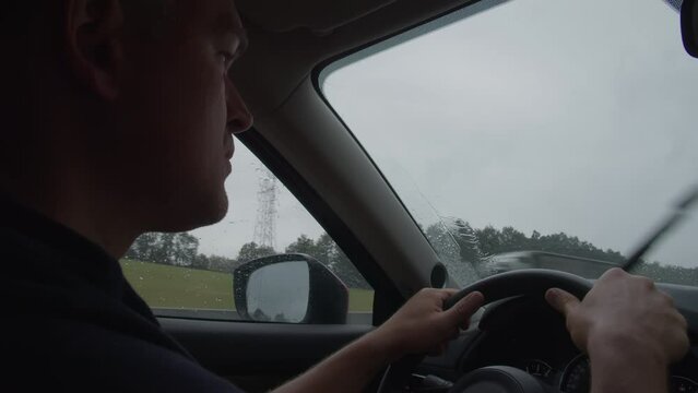 Profile of a man driving a car, focusing intently on the road during a rain shower, with the windshield wipers in motion and raindrops visible on the side window