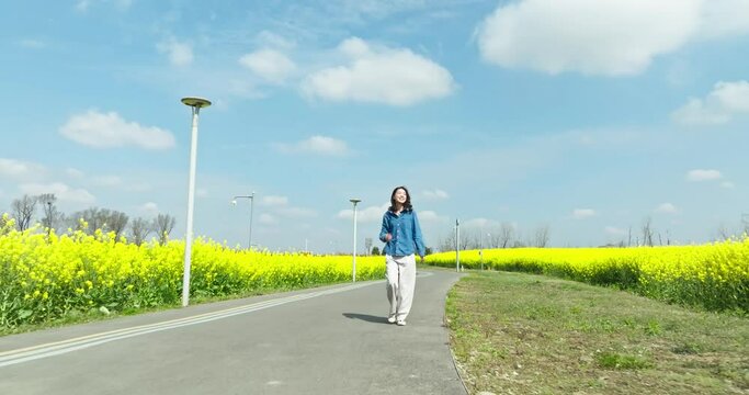 happy young woman walking on the countryside road at spring sunny day with blooming oilseed flower field