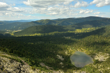 A view from the top to a beautiful lake surrounded by mountain taiga on a sunny summer day.