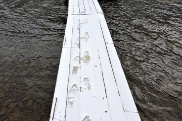 Human footprints on a snow-covered wooden platform above the calm water of a mountain lake.