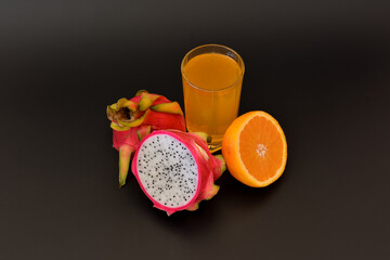 Mixed fruit juice in a glass on a black background, next to a ripe cut orange and pitaya.