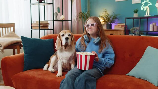 Excited young girl in 3D glasses eating popcorn and watching interesting TV serial, sport game, film, online movie content. Preteen child kid sits beside beagle dog on couch in living room at home.