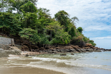 A steep rocky seashore overgrown with tropical vegetation next to a sandy beach at low tide. In the...