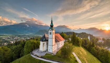 skofja loka slovenia aerial panoramic view of the beautiful hilltop church of sveti tomaz saint thomas with amazing golden sunset and the julian alps at background at summer time - obrazy, fototapety, plakaty