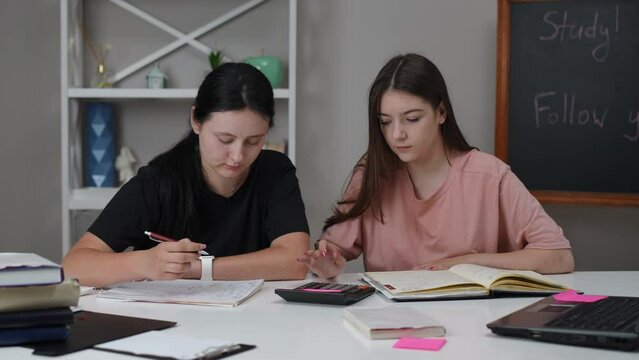 Two female students are preparing for an exam, sitting at a table in the living room. One girl rereads her lectures in a notebook and counts on a calculator, while the other writes with a pen in a