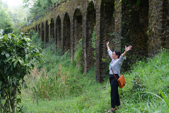 Side view: A woman standing next to a series of pillars of an old bridge while raising her hands