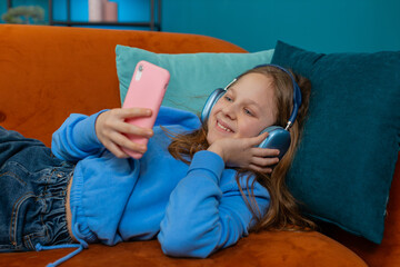 Happy Caucasian girl child in wireless headphones relaxing lying on couch at home listening...