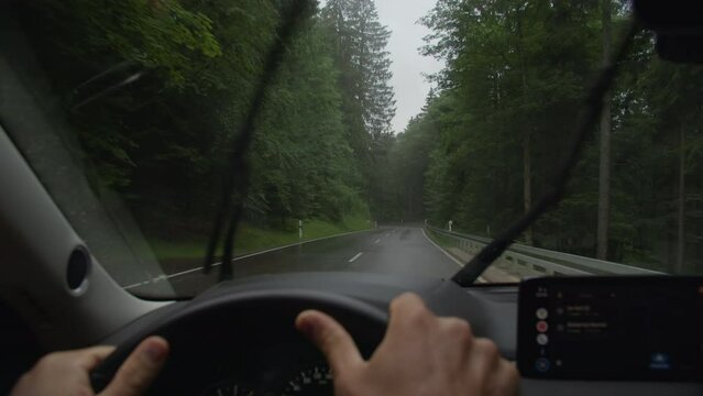 An inside view of a car driving on a wet forest road, with the windshield wipers turned on, past green trees and a road sign ahead