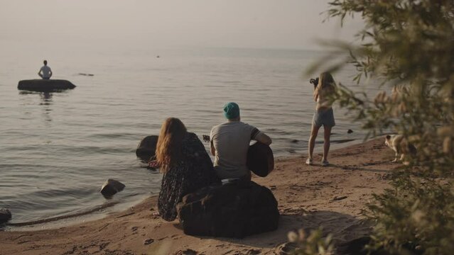 A group of hipster people spend holidays together at sea. The guy playing guitar to his friends at the backdrop of sunset. A girl photographs a guy in a river. isolation together, vacation concept.