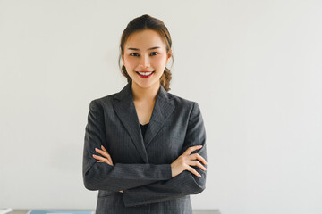 Professional Asian businesswoman smiling confidently with her arms crossed, wearing a grey suit in office.