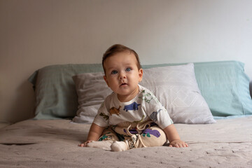 Bebé de ojos azules y cabello castaño sentado en una cama en su habitación usando polo con dibujos, sentado defrente, pared blanca