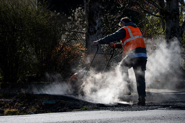 Construction worker using a plate compactor to finish the fresh asphalt at the end of a sidewalk while early morning sun highlights the steam and smoke, road re-paving project
 - obrazy, fototapety, plakaty