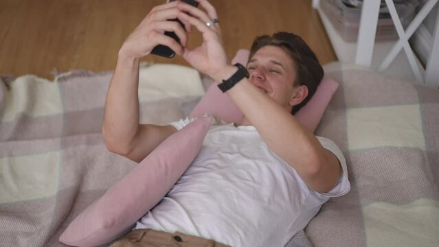 The guy records a video message on the camera of his mobile phone in his hands, lying on a wooden floor on a blanket with pillows under his head. The guy smiles, waves and gestures with his free hand.