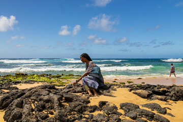 an African American woman wearing sunglasses with long sister locks relaxing at Sandy Beach with rocks covered in green algae, blue ocean water, crashing waves blue sky and clouds in Honolulu Hawaii