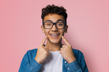 Dental dent care ad concept image - black сurly haired funny young man in metal braces wear eye glasses, show point white teeth smile. Isolated rose pink studio wall background. Positive optimistic.