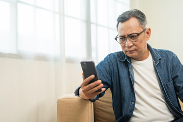 Depressed Asian mature old man using smartphone serious thinking on sofa in living room. Portrait...