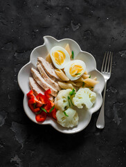 Healthy lunch - baked chicken breast, egg, sweet pepper and boiled cauliflower on a dark background, top view