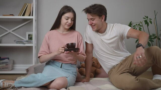 A girl reads a book while sitting on a plaid on the floor. The guy brought a coffee cup and saucer and gave it to the girl. The guy sat down with the girl on the blanket and they started talking