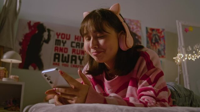 Teenage girl listens to music in headphones and scrolls social media using mobile phone lying on the bed in the cozy bedroom. Asian girl spending pastime and having fun at home. Lifestyle concept.