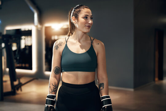 Fit young woman wearing a tank top and boxing gloves smiling while standing in a gym during a self defence and workout session