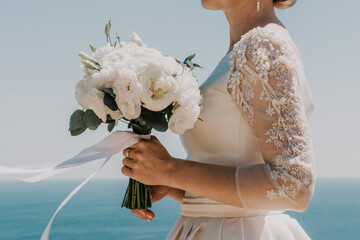 bride is holding a bouquet of white flowers. The bouquet is large and has a lot of flowers in it. The woman is wearing a white dress and a gold ring on her finger. Scene is elegant and romantic.