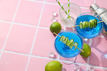 Blue cocktail with cucumber slice in glasses, shaker, limes and ice cubes on pink background, space for text