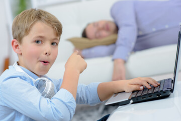 Young boy on laptop pointing to sleeping adult - obrazy, fototapety, plakaty