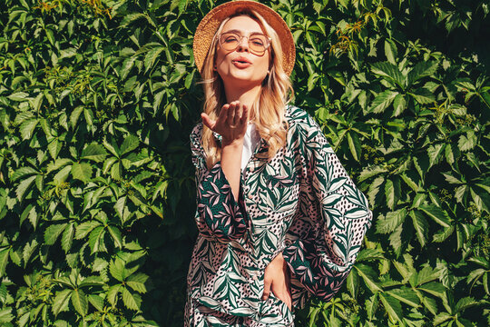 Young beautiful smiling blond hipster woman in trendy summer hippie costume clothes. Carefree female posing in street. Positive model. Cheerful and happy in hat near green bushes. Give air kiss