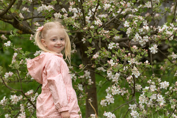 Portrait of happy beautiful blonde girl wears in pink jacket stands in spring garden near flowering tree. Smiling girl