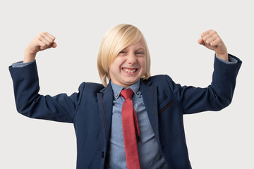Portrait of a happy little boy with blond hair wearing a blue jacket and red tie, raising his arms in triumph, celebrating success or victory