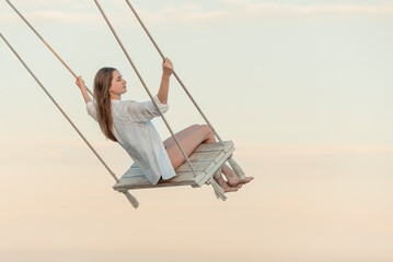 Beautiful young girl in white clothes pushes in swing on sky background. Beautiful soft sunset light. Pretty girl hovers in sky.