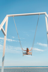 Girl with long hair on rope swing against on sea and blue sky background. Concept of freedom and child carelessness. Vertical frame.