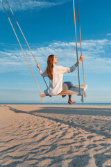 Blond young woman with long hair and white shirt sits on swing above sea. Magic sunset lights. Back view