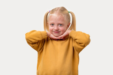 Portrait of a little girl with blond hair in a yellow sweatshirt holds her hands to her chin.