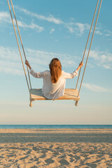 Blond young woman with long hair and white shirt sits on swing above sea. Vertical frame. Yellow magic sunset lights. Back view