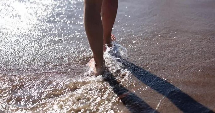 Slender female legs and feet walking on the waves of sea water on sandy beach. Beautiful woman walking on the sea surf