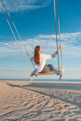Happy girl have fun swinging high in mid air. Young woman in white shirt on rope swing on blue sky and sea background