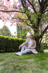 Young woman sitting on lawn in blossoming park, warm spring day. Attractive girl on rest. Vertical frame.