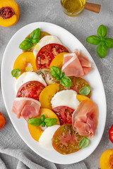 Salad of sliced multi-colored tomatoes, fresh peaches, mozzarella, prosciutto and basil on a gray concrete background. Delicious summer salad.