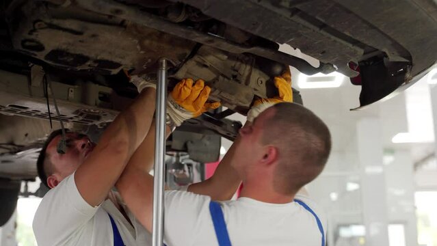 Auto mechanics work on vehicle transmission removal in garage. Car repair, teamwork, undercar service procedure. Technicians dismantle gearbox, manual mechanic work. Auto shop, maintenance job.