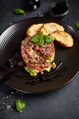 Tuna and avocado tartare with sesame seeds, capers and egg yolk on a dark ceramic plate with baguette bread crouton chips