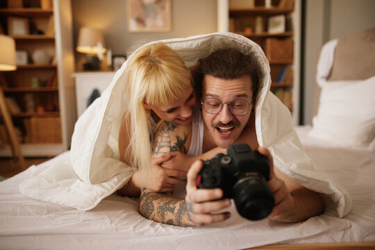 Tattooed man photographing his wife with a camera in bed