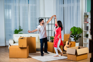 Happy Indian young couple dancing in modern living room with cardboard boxes with belongings.