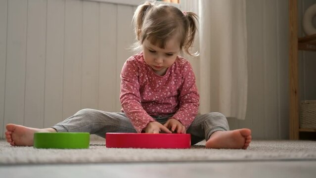 Kid girl play toys at home, kindergarten or nursery. Baby child playing playthings cubes and wood puzzle, pyramid sitting on the floor. Happy preschool education indoor. 3 years old little toddler.